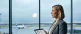 Suomalainen nainen seisoo toimiston ikkunalla jonka takana lentokenttä, tabletti kädessä