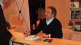 Homme âgé en veste élégante, assis à un bureau de studio, parlant avec énergie et confiance dans un cadre professionnel