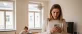 Österreichische Mutter mit Smartphone in Wiener Wohnung, Kind im Hintergrund auf Tablet