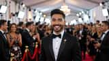 Young Latino comedian in formal black suit at a gala red carpet event with camera flashes
