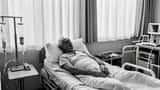 Patient in NHS hospital bed with IV drip, family member providing support
