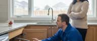 Plumber in blue coveralls explaining a pipe issue to a homeowner in a Canadian kitchen