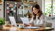 Australian woman reviewing 2026-27 budget documents at a Perth suburban kitchen bench, laptop open to myGov, warm morning light through window