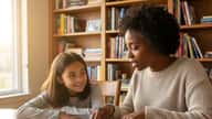 African American English tutor helping a young girl read a book at a home study table in suburban Chicago