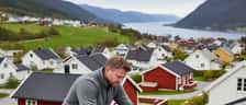Norsk mann monterer solcellepanel på taket av et rødt trehus med fjordlandskap i bakgrunnen