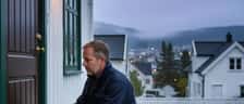 Låsesmed kneler ved inngangsdøren til et hvitt norsk trehus og arbeider med profesjonelt verktøy i skumringen