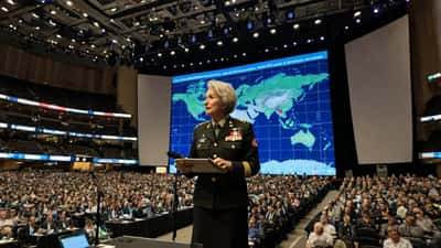Australian military general speaking at the Space Symposium 2026, with satellite and cyber security imagery projected on screens behind her