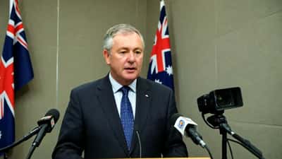 Australian politician addressing press conference with Australian flag background in Canberra
