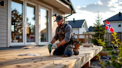 Mann i arbeidsklær monterer terrasseplanker med drill foran et norsk hus om sommeren