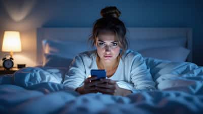 Ung kvinna vaken i mörkt sovrum, ansiktet upplyst av mobilens sken med nyhetsnotiser