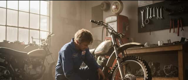 Australian motorbike mechanic in overalls kneeling beside a motorcycle in a sunlit workshop, checking tyre pressure