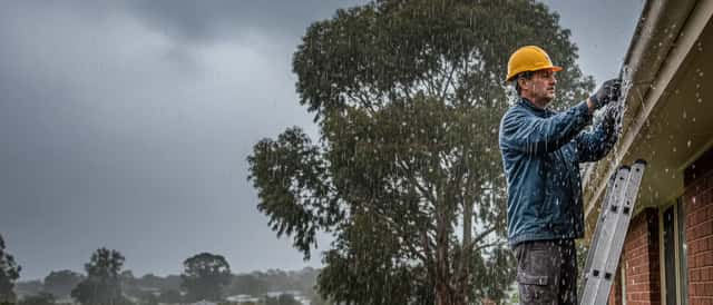 Tradesperson inspecting roof gutters in heavy rain, Australian suburban home, overcast sky