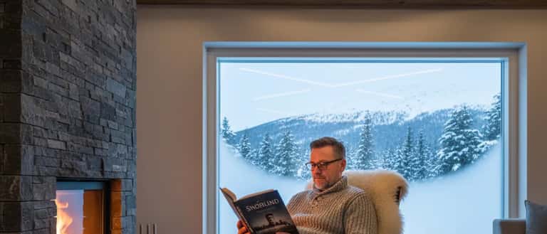 Mann leser krimroman ved peisen i norsk fjellhytte om vinteren