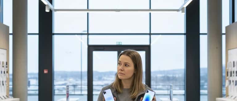 Nordisk kvinne sammenligner to smarttelefoner i en lys norsk elektronikkbutikk