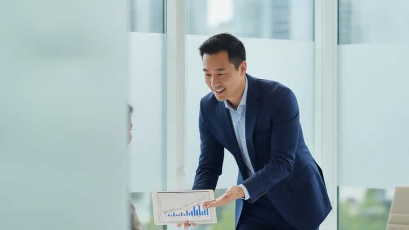 East Asian Canadian financial advisor in a Vancouver glass-fronted office showing a portfolio growth chart on a tablet to a client across a clean desk