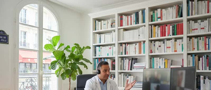 Médico brasileiro em teleconsulta por videochamada, gesticulando enquanto explica orientação ao paciente