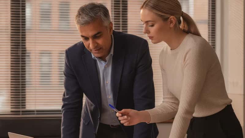 An HR director in a Manchester office explaining pension auto-enrolment paperwork to a younger colleague at a conference table, illustrating the new communication obligations employers face under the 2026 expansion