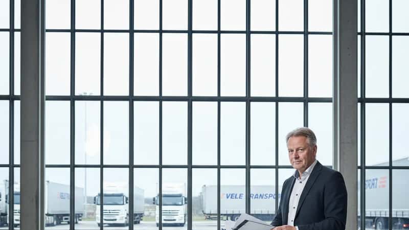 HR-direktør i Vejle transportvirksomhed gennemgår personalekontrakter på stående arbejdsstation med fragtbiler synlige gennem vinduer