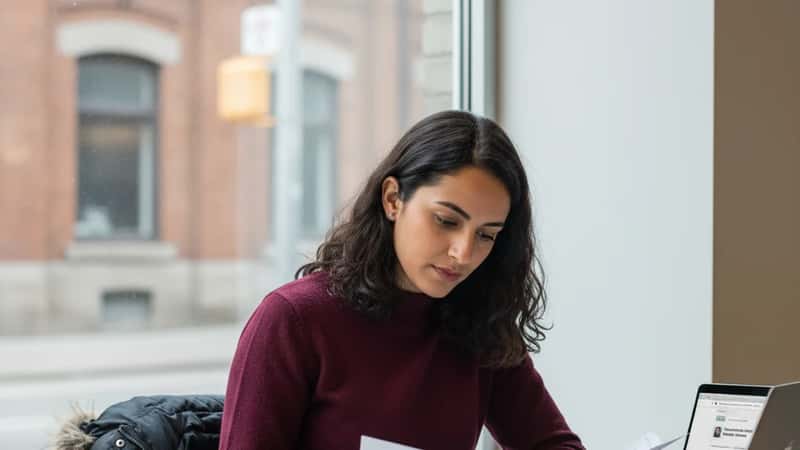 Jeune femme d'origine maghrébine étudiant ses documents d'immigration dans un café d'Ottawa, ordinateur portable ouvert sur le site d'IRCC