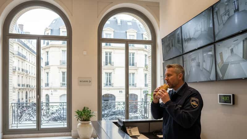 Security guard eating a sandwich at a front desk in a New York office building lobby while monitoring security screens, on-duty during break