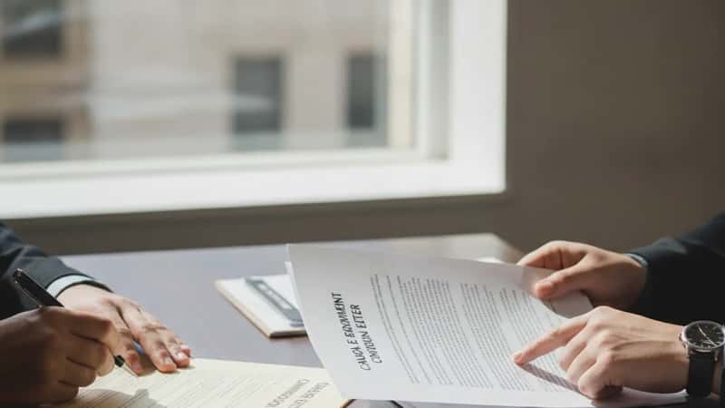 Two professionals across a meeting table in a Melbourne law office, one signing a casual employment conversion letter, the other reviewing the employer agreement, natural daylight, decisive professional moment