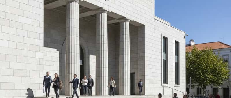 Fachada de tribunal português com colunas de pedra e símbolo da balança da justiça, cidadãos a subir as escadas num dia de sol