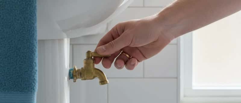 Homeowner turning a brass stopcock valve under a bathroom sink in a British home