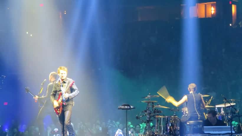 Muse performing on stage at an indoor arena, bright stage lights illuminating the crowd