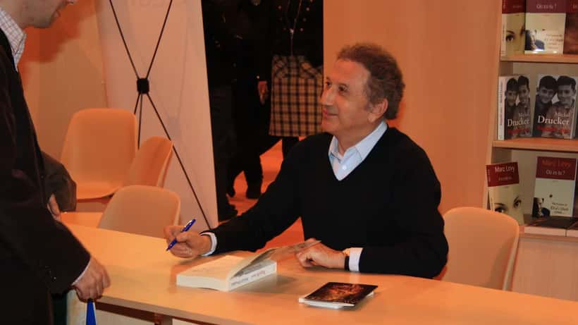 Homme âgé en veste élégante, assis à un bureau de studio, parlant avec énergie et confiance dans un cadre professionnel