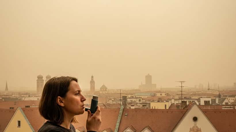 Frau mit Asthma-Inhalator auf Balkon blickt auf gelblichen saharastaubverhangenen Münchner Himmel