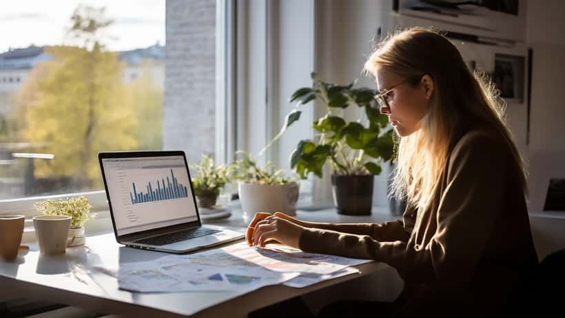 Nuori suomalainen nainen tarkastelee yritystään taloudellisia tietoja kannettavalla tietokoneella kotitoimistossaan Helsingissä