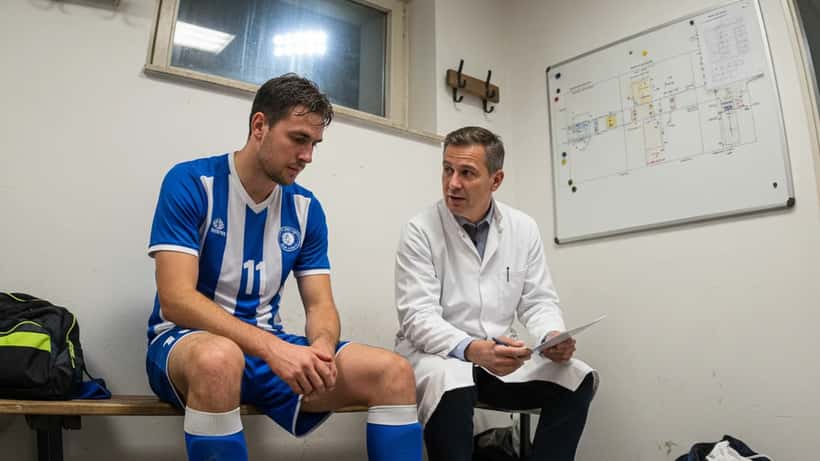 Amateuerfußballer mit Knöchelverletzung beim Gespräch mit Sportarzt im Umkleideraum eines deutschen Fußballvereins