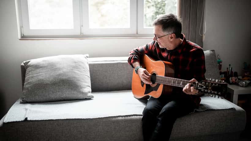 Aelterer Mann spielt Akustikgitarre allein zu Hause, konzentriert auf sein Instrument