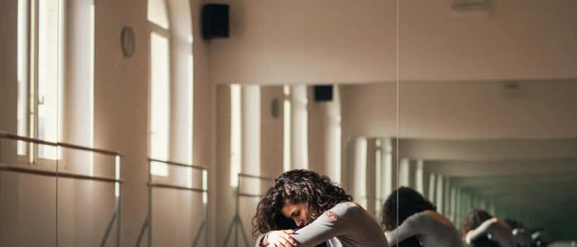 Giovane donna in sala prove di danza, seduta a terra, espressione tesa e stanca