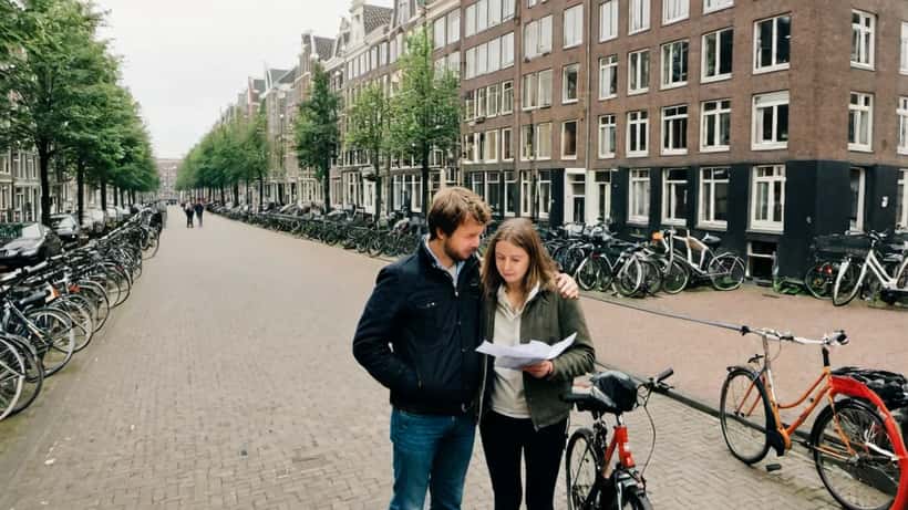 Jong stel bekijkt hypotheekpapieren voor Amsterdams appartement met Te Koop bord na het lezen van Maaike Schoons boek over de woningcrisis