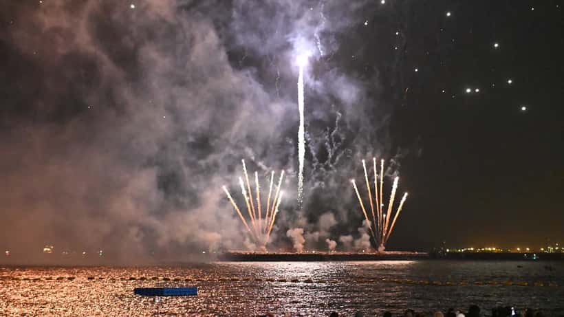 Crowd watching fireworks burst and reflect over calm water at a riverfront festival at night
