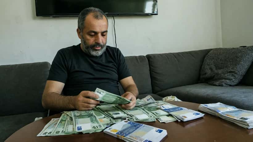 Homem português preocupado a verificar apostas desportivas na Ligue 1 no telemóvel, com notas de euro na mesa