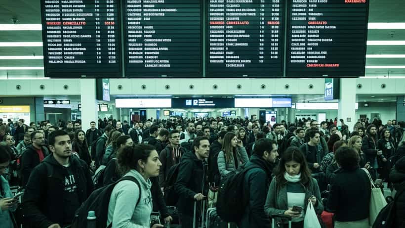Passeggeri frustrati con bagagli attendono in una hall affollata a Roma Fiumicino con tabellone che mostra voli cancellati