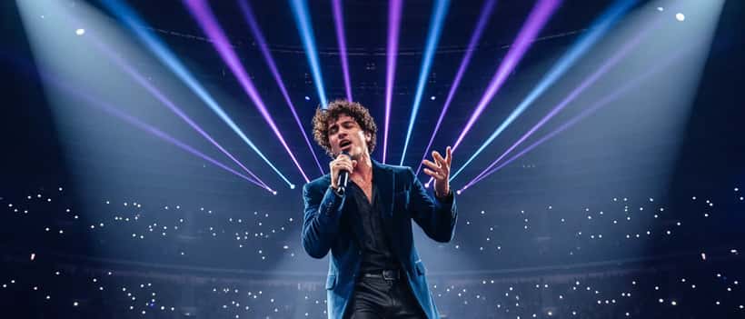 Cantante italiano sul palco di un palazzetto con microfono, luci blu e viola, folla enorme sullo sfondo