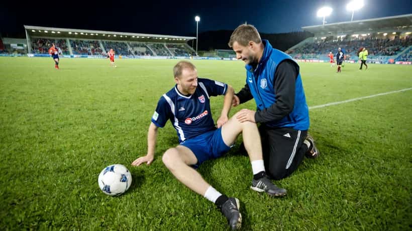 Amatørfotballspiller sitter på sidelinjen og holder seg til kneet mens en idrettslege undersøker ham under en kamp