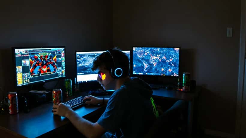 Joven adulto jugando videojuegos de superhéroes en PC en una habitación oscura con múltiples pantallas encendidas, latas de bebida energética en el escritorio