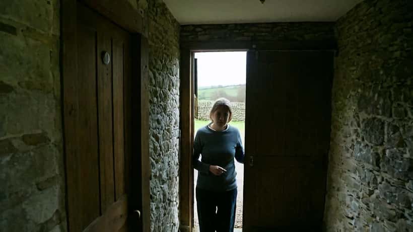 Woman standing at a heavy door in a dimly lit hallway, symbolising isolation and coercive control in a closed community