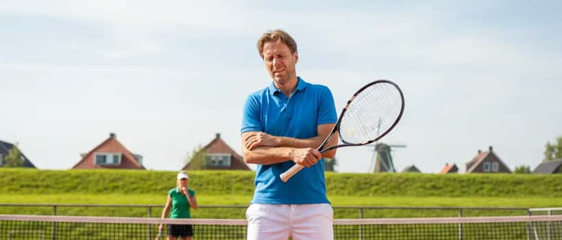 Een Nederlandse recreatieve tennisser op een buitenbaan die zijn elleboog vasthoudt van pijn