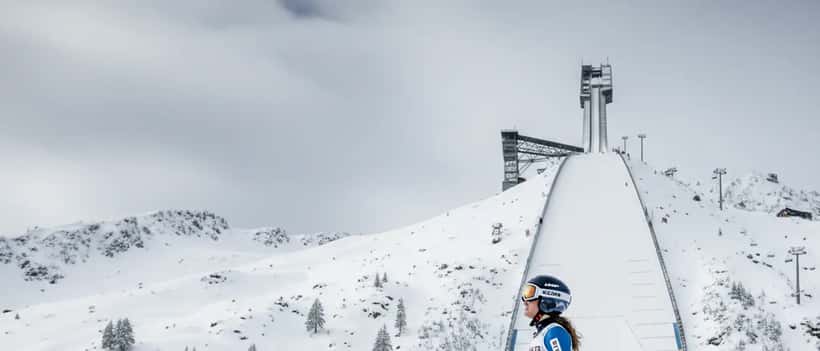 Skifahrerin in Profiausrüstung blickt auf eine große Skiflugschanze in Winterlandschaft