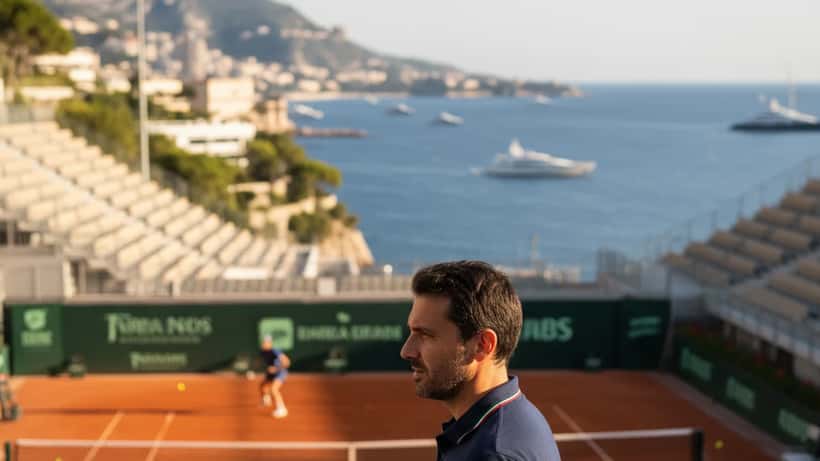 Allenatore di tennis su campo in terra rossa a Monte Carlo con appunti e giocatore sullo sfondo