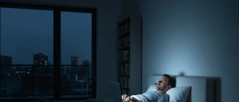 Person liegt wach im dunklen Schlafzimmer, Bildschirmleuchten im Gesicht, Uhr zeigt 1 Uhr nachts