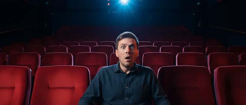 British man gripping cinema seat armrests with wide eyes watching a scary film