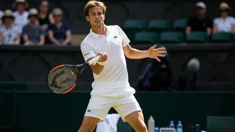Dansk tennisspiller serverer på grønt baneunderlag med sportsmedicinsk udstyr ved banen