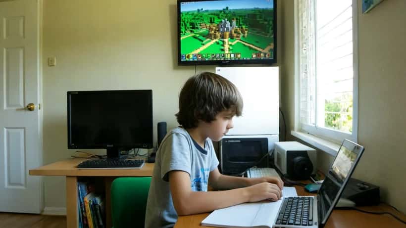 Enfant français jouant à Minecraft sur ordinateur, cahiers de cours posés sur le bureau à côté