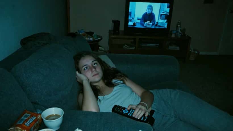 Young woman watching TV alone at night on a couch, exhausted, phone and remote nearby
